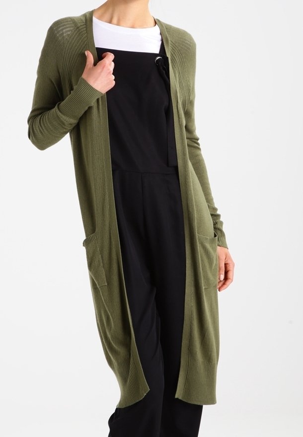 Personne portant un long cardigan vert olive ouvert sur le devant, par-dessus un haut blanc et une salopette noire, les mains près des poches et de la poitrine.