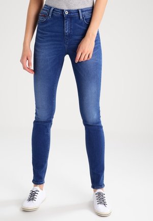 Frau trägt eng anliegende blaue Jeans, graues Oberteil und weiße Sneaker, steht vor einem einfarbigen hellen Hintergrund.