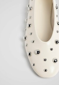 Off-white flad sko med rund tå, dekoreret med sølvfarvede nitter i forskellige størrelser, der er jævnt fordelt på den øverste overflade.