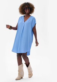 Jasnoniebieska sukienka z krótkim rękawem, dekoltem w kształcie litery V i luźnym krojem, z bocznymi rozcięciami. W zestawieniu z beżowymi kowbojkami i dużymi okularami przeciwsłonecznymi.