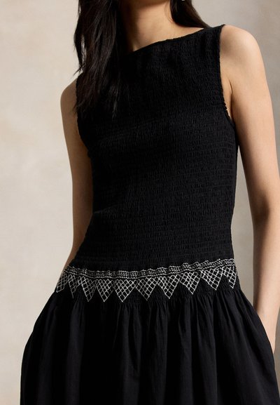 Hihaton musta mekko, jossa teksturoitu bodi ja valkoinen geometrinen kirjonta vyötäröllä, päällä tummahiuksinen henkilö.