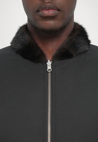 Čierna bunda s lesklým strieborným zipsom a mäkkým, tmavým kožúškom na golieri. Záber zblízka zdôrazňuje textúru látky a detaily goliera.