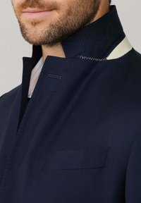 Granatowa kurtka z materiału, z wysokim kołnierzem, z białymi wykończeniami, przednią kieszenią i kontrastowym haftem. Gładka tekstura i dopasowany krój.