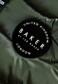 Patch circolare nera con testo bianco su un tessuto imbottito verde oliva. Il testo include "BAKER," "REGNO UNITO," e "LONDRA." Texture liscia.