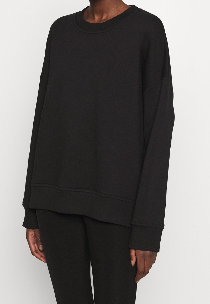 Personne portant un sweat-shirt noir ample à manches longues avec poignets et ourlet côtelés, assorti à un pantalon noir sur un fond uni.