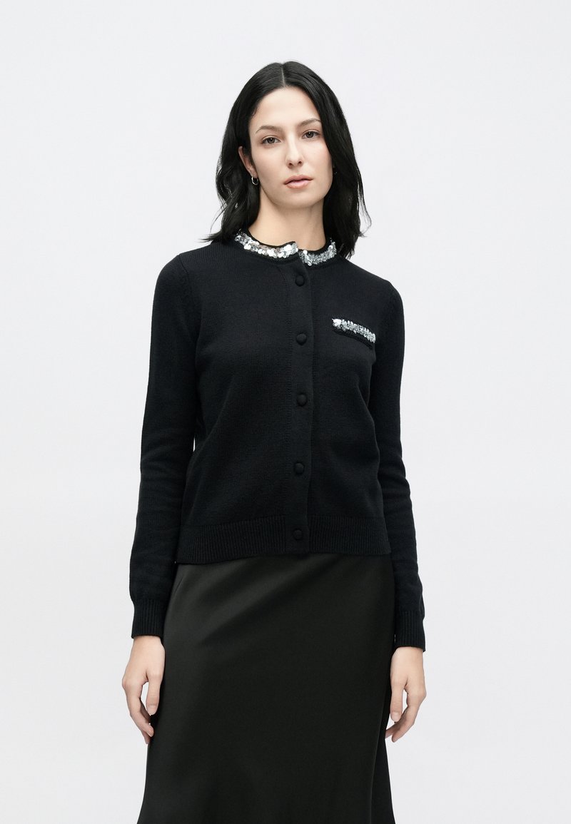 Black cardigan made of soft knit fabric, featuring a sequin-embellished collar and pocket, with a fitted design and button closure.