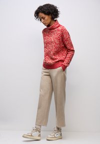Suéter de punto de cuello alto con estampado rojo y crema, combinado con pantalones beige de corte ajustado y zapatillas ligeras. Materiales texturizados visibles.