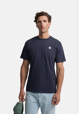 Hombre con cabello rizado que lleva una camiseta azul marino y vaqueros azul claro, sosteniendo un sombrero verde en su mano izquierda contra un fondo liso.
