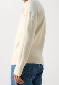 Béžový svetr se žebrovanou texturou, přiléhavým střihem a kapsou na zip na rukávu. Kombinován s modrými džíny z denimu.