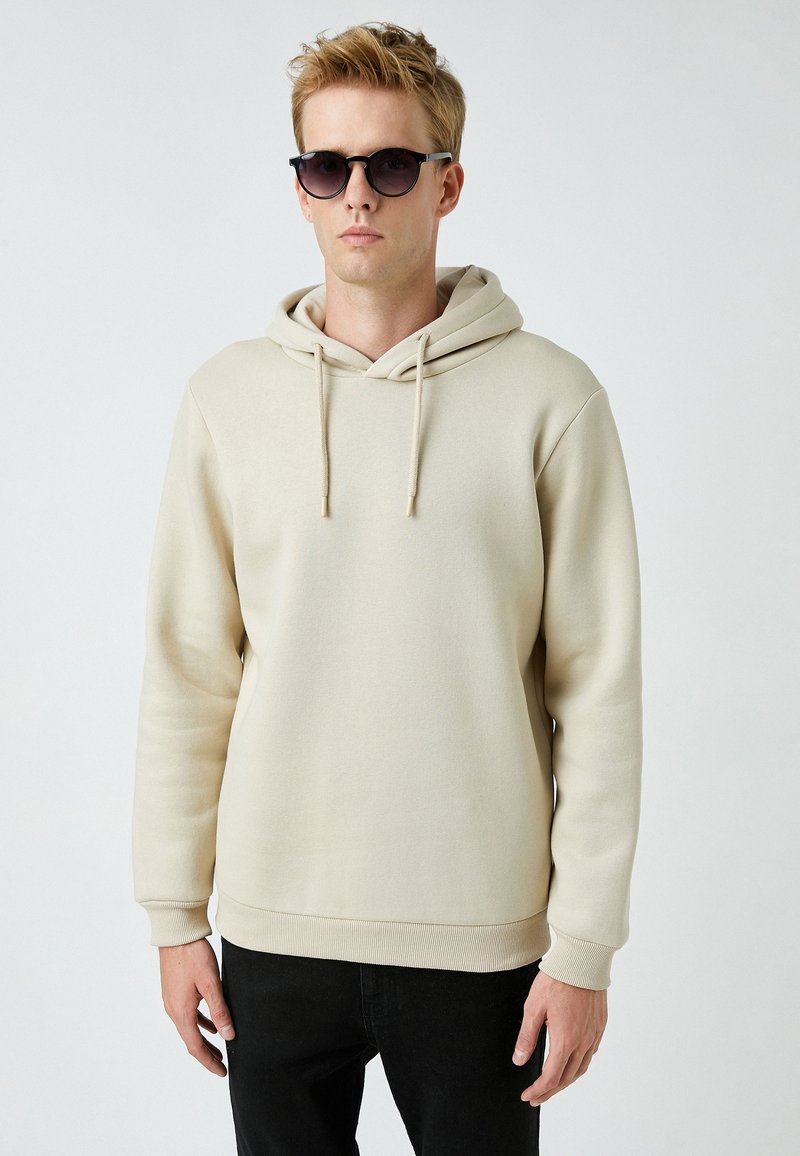 Sudadera beige con bolsillo canguro y capucha con cordón, hecha de un tejido suave. El modelo lleva pantalones oscuros y gafas de sol negras.