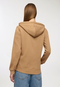 Felpa marrone con cappuccio e polsini a costine, abbinata a jeans azzurri a vita alta. Il tessuto sembra morbido e informale.