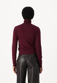 Osoba z krótkimi kręconymi włosami, ubrana w dopasowany bordowy sweter z golfem i czarne skórzane spodnie, widziana z tyłu.