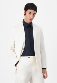 Heren crèmekleurig pak van gladde stof, met een op maat gemaakte blazer en slim-fit broek, gecombineerd met een diep marineblauw overhemd met knopen.