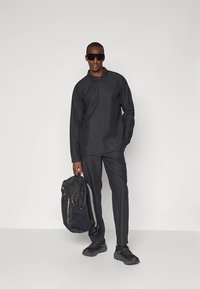 Mężczyzna ubrany w całość na czarno: długą koszulę, spodnie, czarne trampki, okulary przeciwsłoneczne, trzymający czarny plecak za jeden pasek.