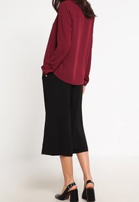 Blouse bordeaux à manches longues avec un ourlet arrondi, associée à un pantalon culotte noir. Des chaussures à talons noires avec des bretelles chevilles complètent la tenue.