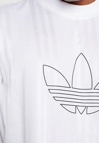 Bílý sportovní tričko se svislými pruhy a černým logem Adidas trefoil ve středu. Hladký povrch, kulatý výstřih.