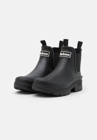 Bottes noires en caoutchouc à cheville avec des panneaux latéraux élastiques et des languettes, présentant une semelle texturée et un logo de marque blanc prominent.