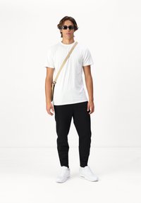 Witte T-shirt en zwarte broek gedragen door iemand. Accessoires omvatten zwarte zonnebril en een beige schoudertas. Witte sneakers maken de look compleet.
