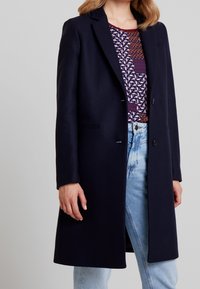 Abrigo de lana azul marino con un corte entallado, solapas muesca, dos botones frontales y bolsillos laterales, llevado sobre una blusa de estampado y jeans.