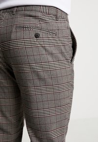 Šedé kárované kalhoty s kombinací černých a vínových proužků. Mají knoflíkový detail na zadní kapse a měkkou, texturovanou látku.