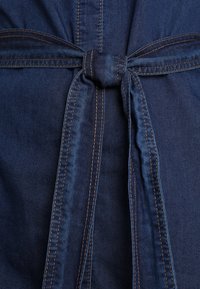 Mørkeblå denim bælte bundet i en simpel knude med synlige brune sømme på stofbaggrund.