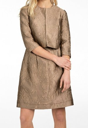 Femme portant une robe taupe texturée et une veste courte assortie, debout, les bras croisés à la taille.