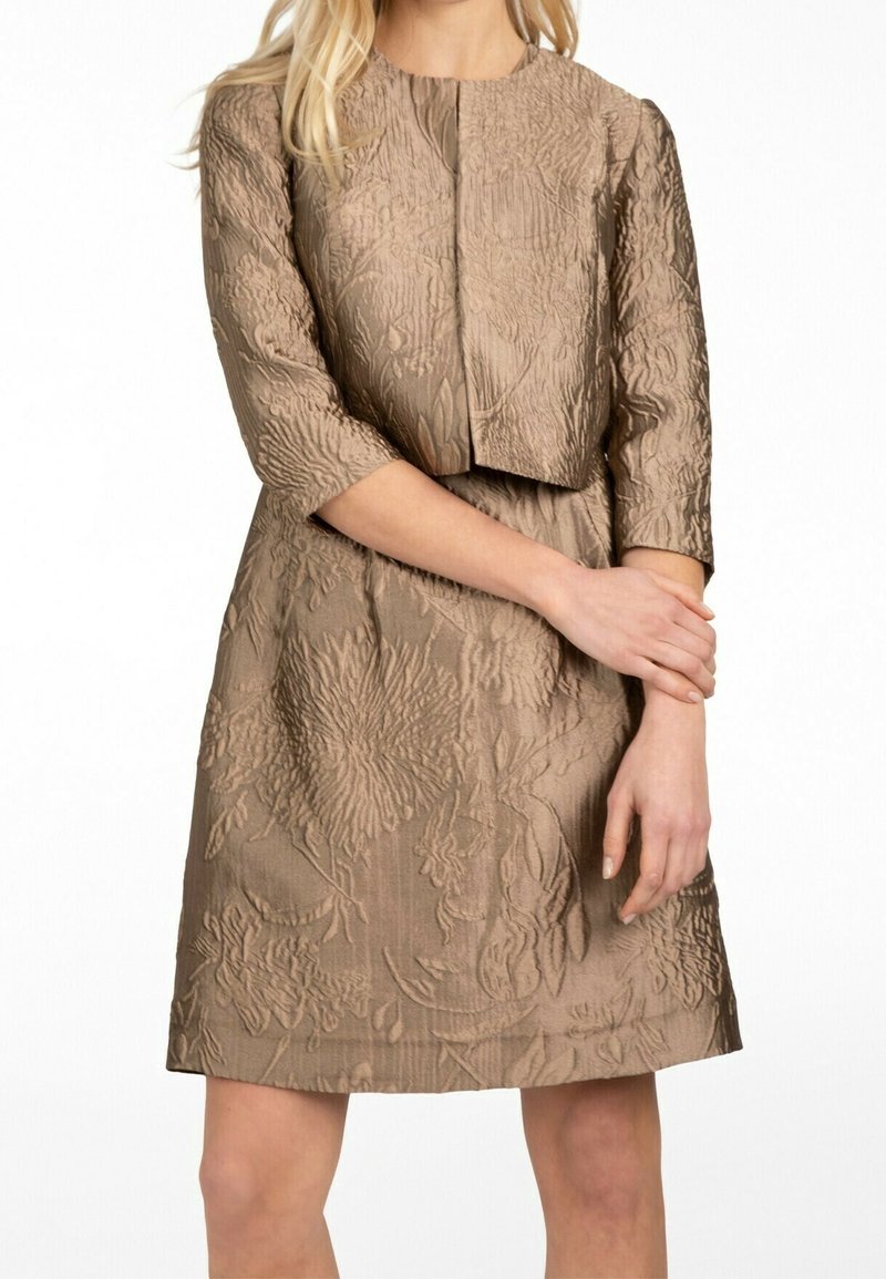Femme portant une robe taupe texturée et une veste courte assortie, debout, les bras croisés à la taille.