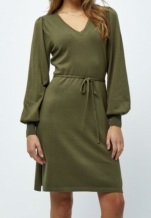 Modèle portant une robe vert olive jusqu’aux genoux, avec un col en V, des manches longues et une ceinture nouée à la taille.