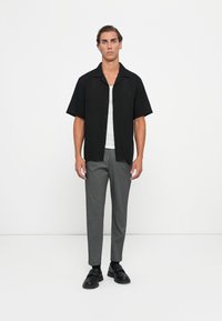 Camicetta nera a maniche corte con bottoni sopra una canotta bianca; pantaloni grigi su misura; scarpe nere. Design minimalista con linee pulite.