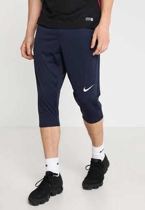 Mężczyzna w granatowych spodniach trzy czwarte marki Nike, czarnych butach Nike, białych skarpetach Nike i czarnej koszulce, stojący na białej podłodze.