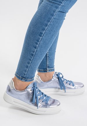 Person wearing blue jeans and metallic silver sneakers with blue laces, standing on a white floor.