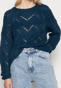 Stickad mörk teal tröja med chevronmönster, lös passform, ribbad mudd och rundad halsringning, ihop med högmidjade ljusblå jeans.