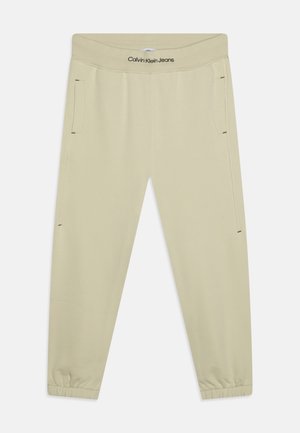 Beżowe casualowe spodnie Calvin Klein Jeans z elastycznymi mankietami, bocznymi kieszeniami i logo na pasie, ułożone na płasko na białym tle.