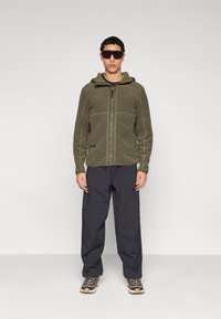 Πράσινο φλις μπουφάν με φερμουάρ μπροστά και κουκούλα, συνδυασμένο με άνετο σκούρο παντελόνι. Το μοντέλο φορά γυαλιά ηλίου και αθλητικά παπούτσια.