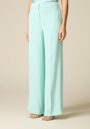 Vrouw draagt lichtblauwe wijde broek met hoge taille en sandalen met open teen op een neutrale achtergrond.