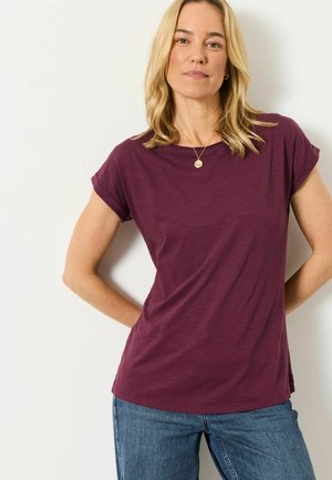 Blonde woman wearing a short-sleeved purple shirt and blue jeans, standing with hands behind back against a white background.