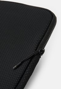 Mala para laptop preta com uma superfície de tecido texturizado e perfurado, fecho suave e um detalhe em cordão para fácil acesso.