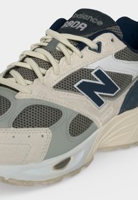 Κοντινό πλάνο ενός γκρι και μπεζ New Balance sneaker με πλέγμα, λευκά κορδόνια και ναυτικό μπλε λογότυπο "N" στο πλάι.