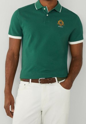 Polo vert avec un col blanc et des accents blancs sur les manches. Logo brodé sur la poitrine. Porté avec une ceinture marron et un pantalon blanc.