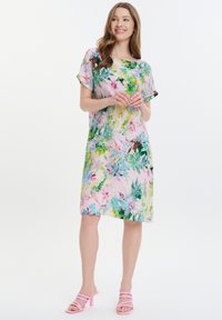 Blumenkleid in Hellrosa mit grünen und blauen tropischen Mustern. Kurzärmeliger Schnitt und lockere Passform, kombiniert mit pinken, hochhackigen Sandalen.