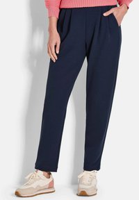 Vrouw in marineblauwe tapse broek, roze sweater en wit-roze sneakers die met één hand in de zak staat tegen een witte achtergrond.