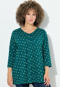Teal langmouwtop met een V-hals, voorzien van kleine gele patronen, een geplooide voorkant en zachte stof. Gecombineerd met donkere jeans.