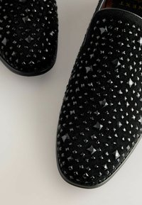 Schwarze Slipper aus strukturiertem Stoff, verziert mit glänzenden, pyramidenförmigen Nieten in unterschiedlichen Größen für dekorative Akzente.