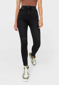 Mujer con pantalones ajustados negros de cintura alta con una rodilla desgastada, combinados con zapatillas blancas y verdes, de pie contra un fondo liso.