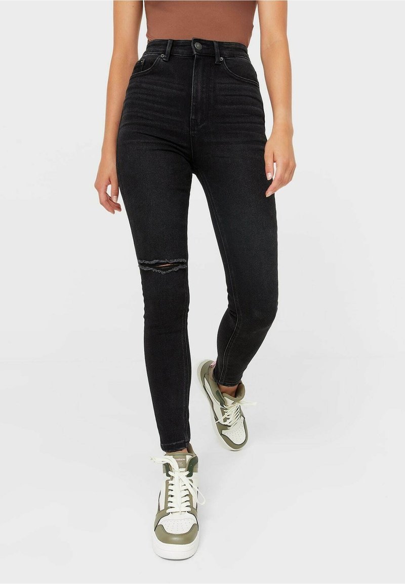 Mujer con pantalones ajustados negros de cintura alta con una rodilla desgastada, combinados con zapatillas blancas y verdes, de pie contra un fondo liso.