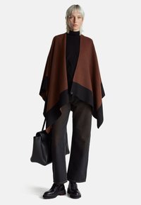 Hnědý a černý pončo s trojúhelníkovým vzorem, nošený přes černý rolák a doplněný černými kalhotami a robustními botami. Součástí je černá kabelka.