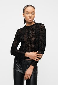 Γυναίκα με πλεξούδες, φορά μαύρο διάφανο τοπ με σχέδιο φίδι και μακρύ μανίκι, καθώς και μαύρο δερμάτινο παντελόνι, ποζάροντας με σταυρωμένα τα χέρια.