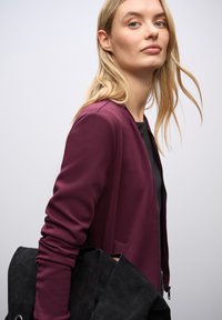 Burgundowa kurtka zapinana na zamek z długimi rękawami i gładką teksturą, w połączeniu z czarną torebką z zamszu. Prosty design, bez wzorów i elementów metalowych.