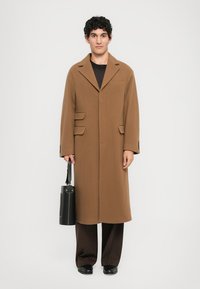 Brown wool coat with a single pocket and three button details, worn over a dark top and brown pants. Black bag held in the left hand.