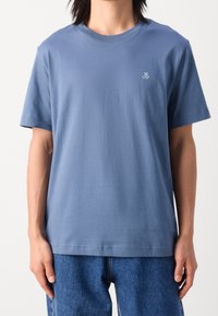 Modré bavlněné tričko s krátkým rukávem, kulatým výstřihem a malým logem na hrudi. Střih je volný. Kombinováno s džínovými kalhotami.